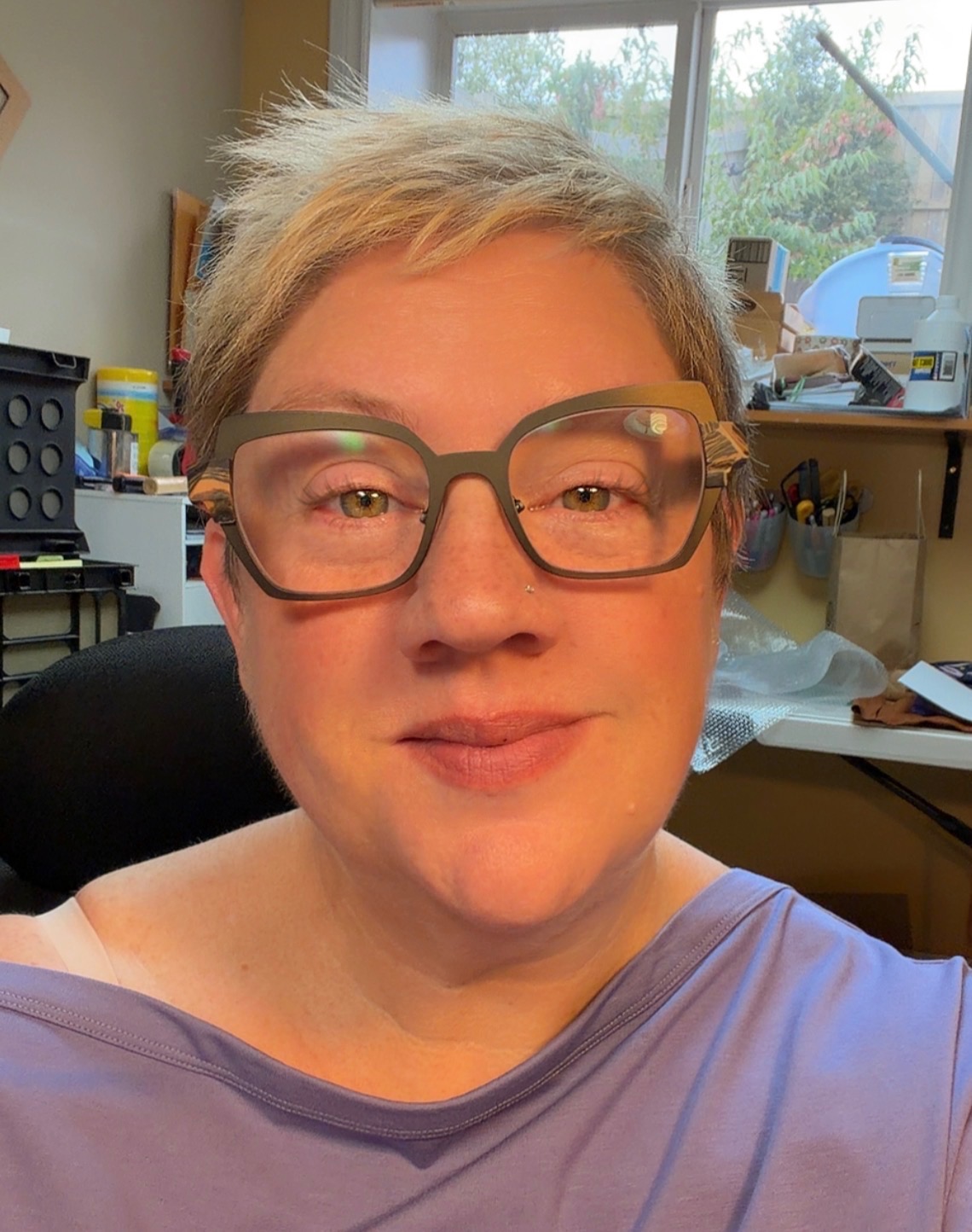 Close-up of a person with short gray hair wearing large glasses, looking at the camera in a bright indoor workspace.